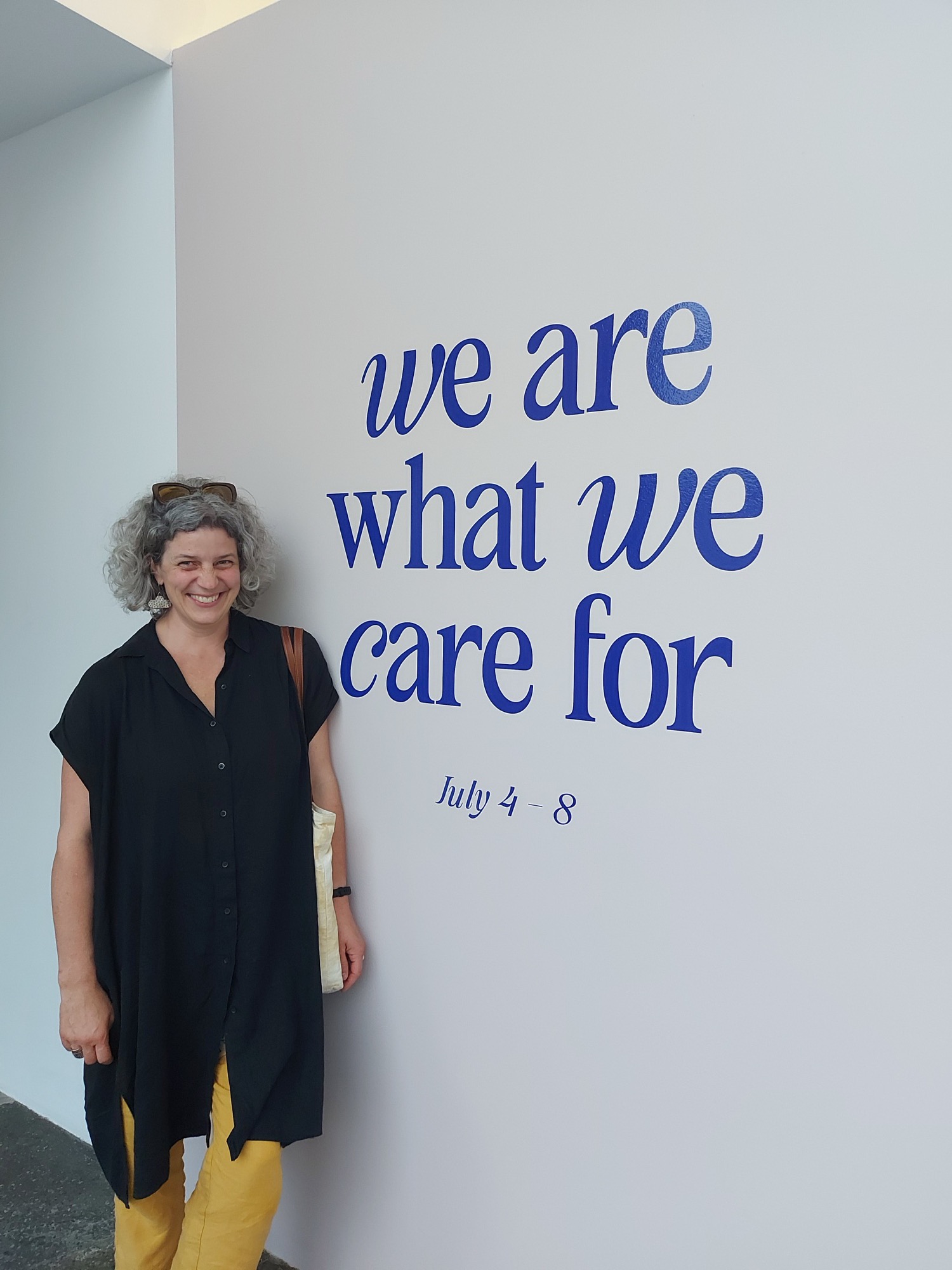 Leslie smiles beside a white wall with the words we are what we care for and July 4 to 8 in large blue letters.