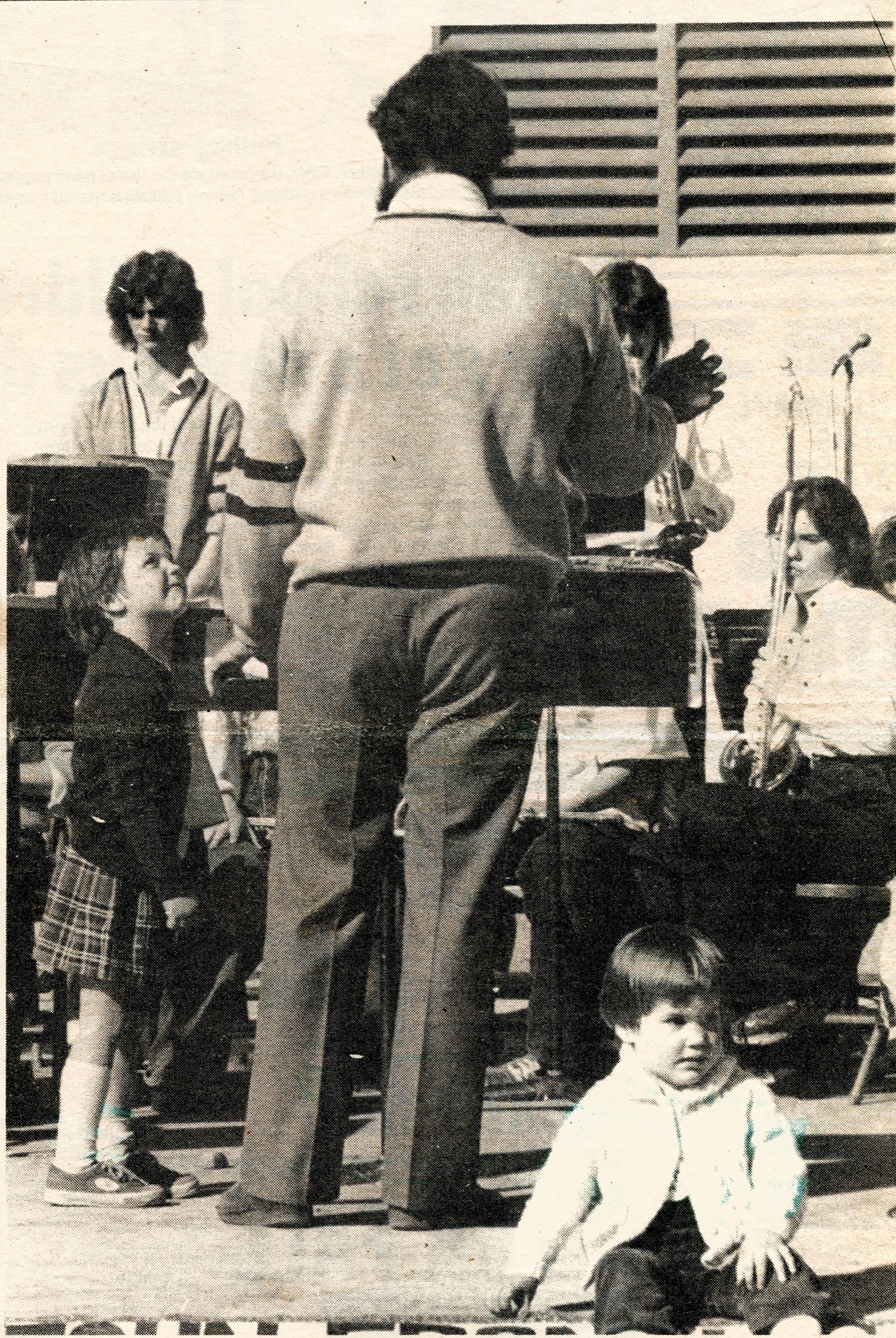 A black-and-white newspaper photo of a band rehearsal. A conductor stands with his back to the camera, one small girl looks up at him, and a toddler sits on the stage in front.