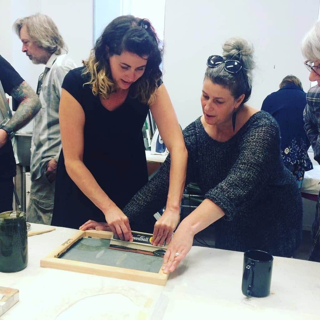 Leslie leans over a print table and guides a student pulling a squeegee across a wooden screen-printing frame.