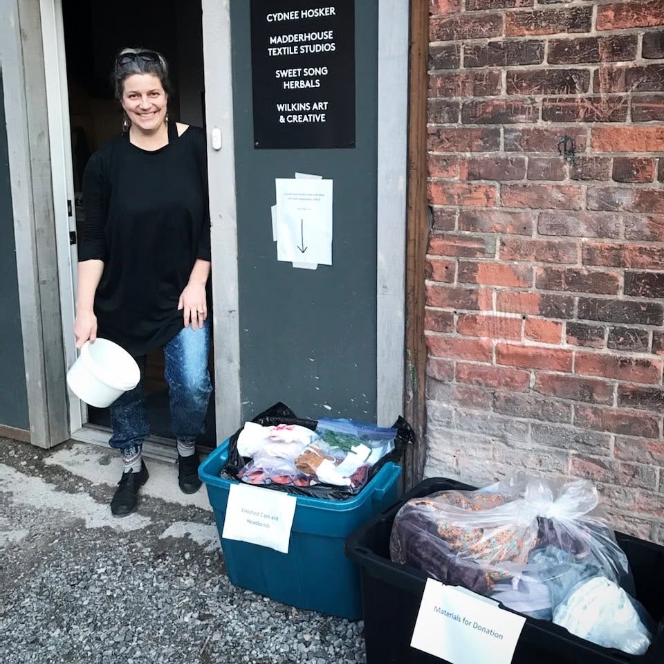 Leslie smiles in a doorway under a sign that lists Madderhouse Textile Studios, holding a white bucket. Two bins beside her are labelled Finished Caps and Headbands and Materials for Donation.