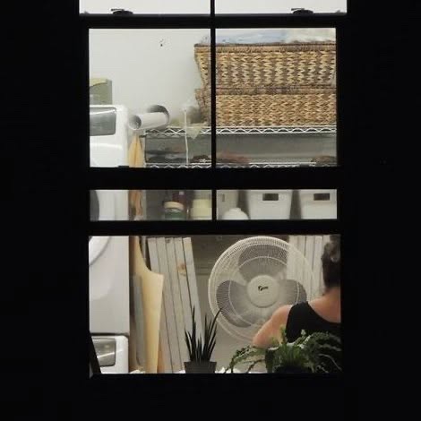A lit window at night looks into the studio, where a woman works beside a fan, a plant, wire shelves and woven baskets.