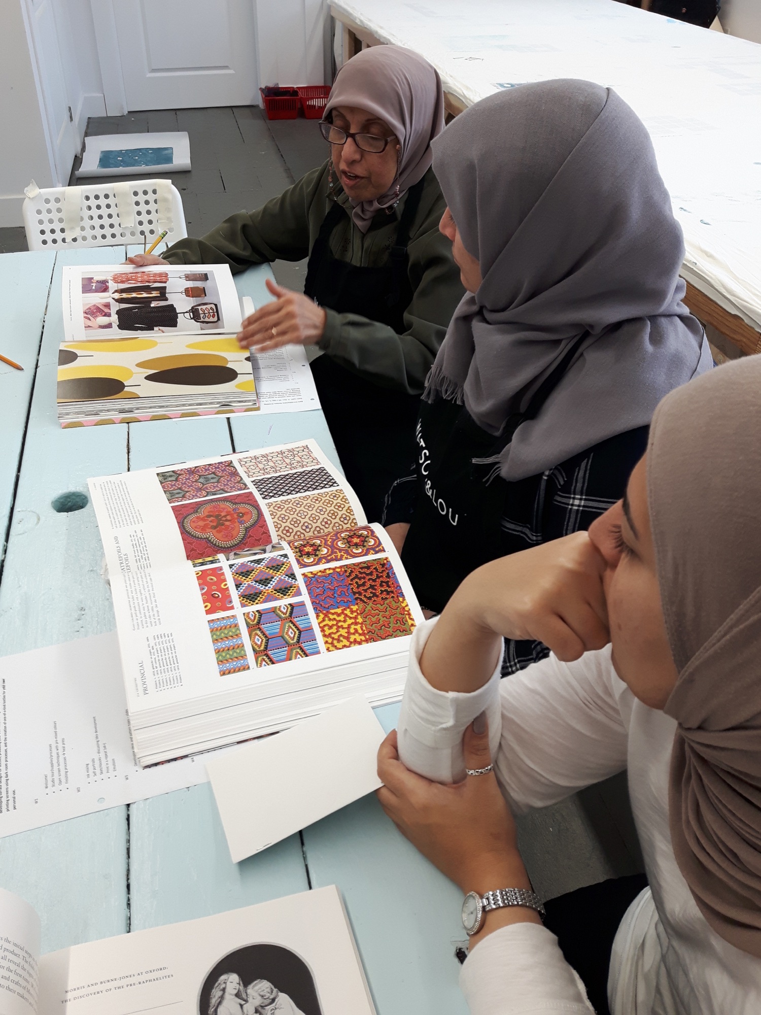 Three women in hijabs sit at a pale blue table with open books of colourful textile patterns. The oldest woman points to a page as she talks.