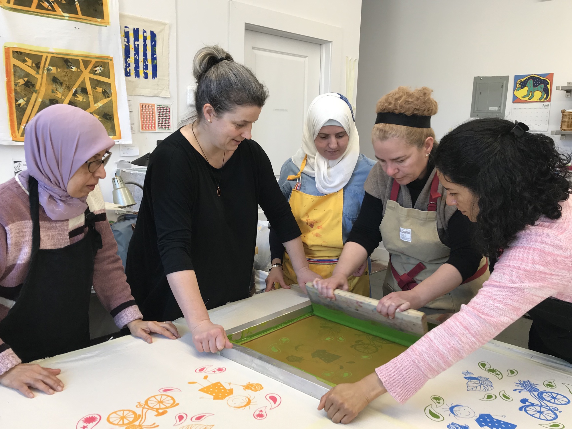 Leslie stands at a long print table as four women gather around a screen. Two of them pull a squeegee across it, and printed bicycles and paisleys in orange and blue cover the fabric.