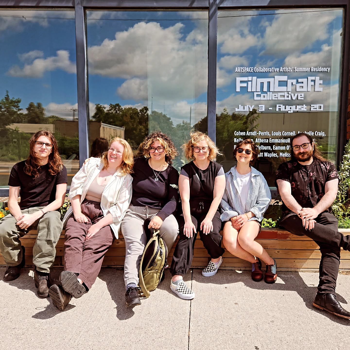 Six artists sit on a wooden bench in front of Artspace’s window, which reads Collaborative Artists’ Summer Residency, FilmCraft Collective, July 3 to August 20.