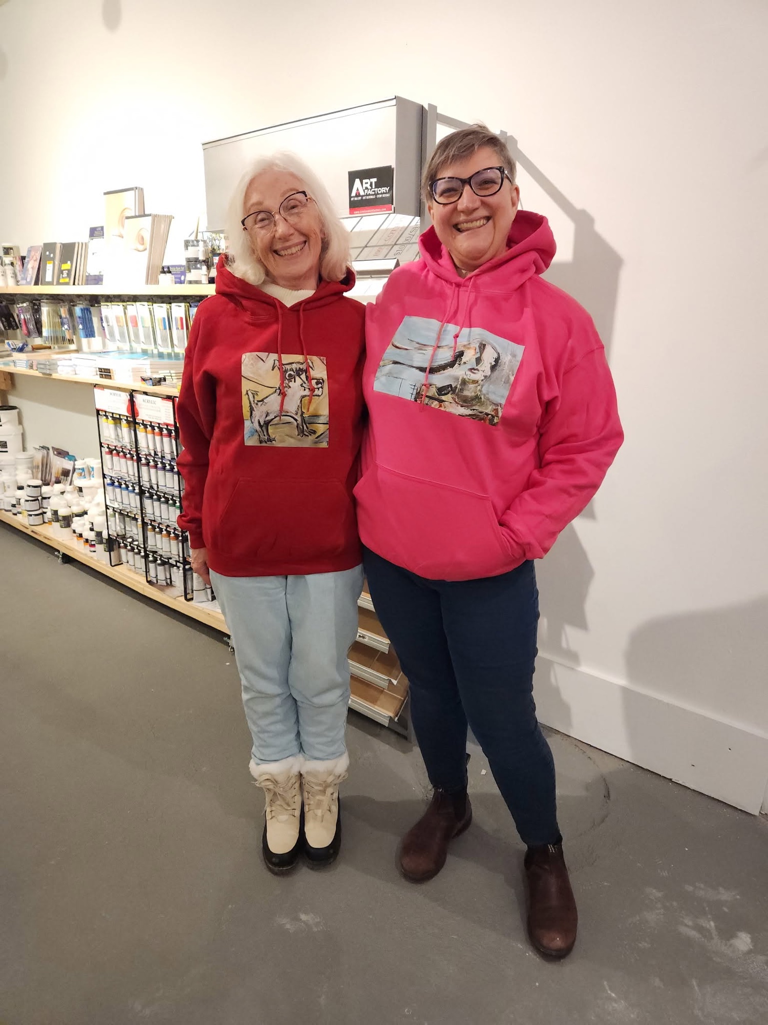 Two smiling women in red and pink hoodies printed with paintings stand in the art-supply shop, beside shelves of brushes, paint tubes and an Art Factory sign.
