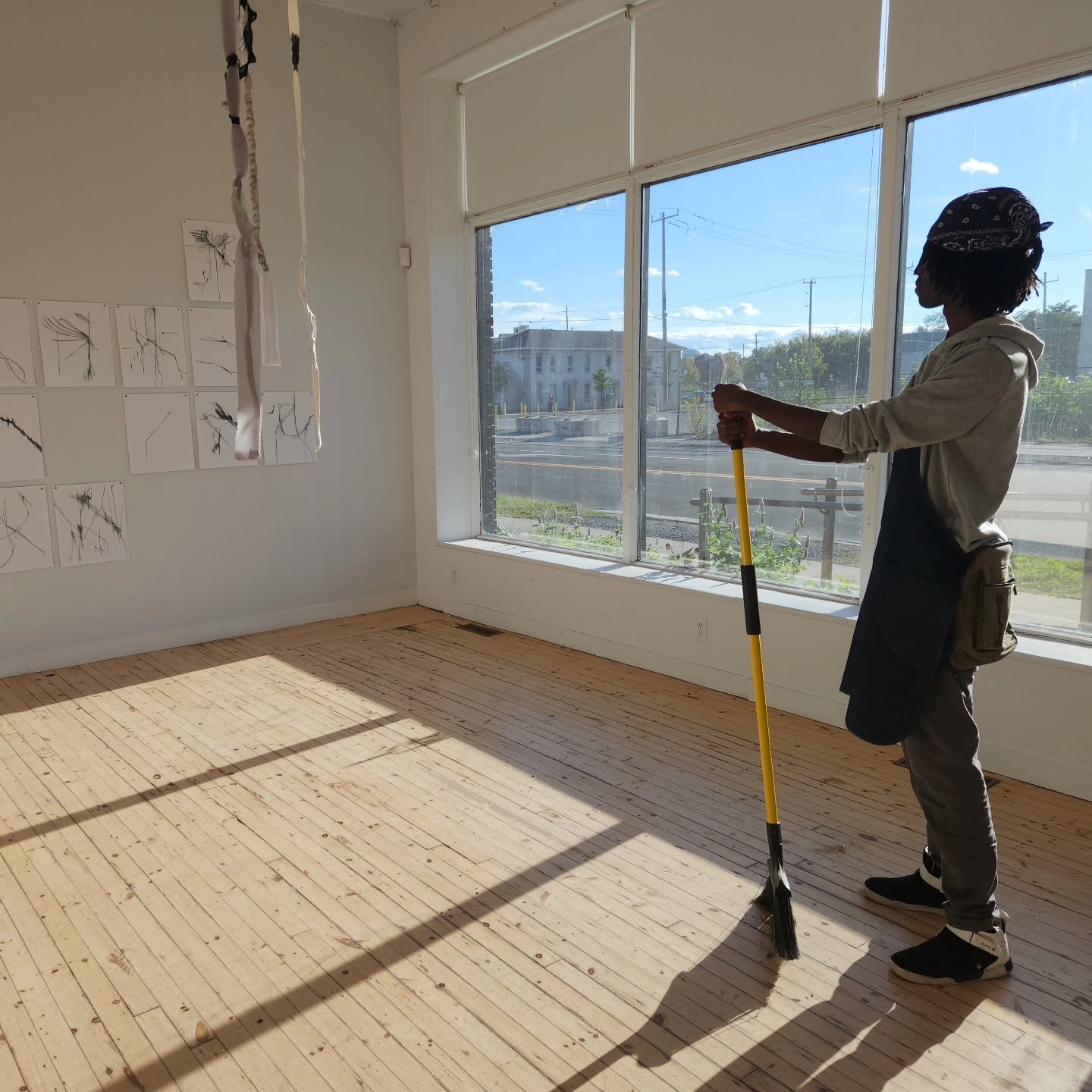 A volunteer in an apron leans on a broom in the sunlit front gallery and looks out the tall street windows. Drawings hang on the wall behind.