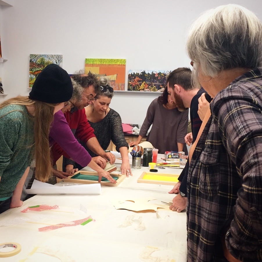 Leslie and a group of adults lean over a long table with small screen-printing frames, tape and cut paper stencils.