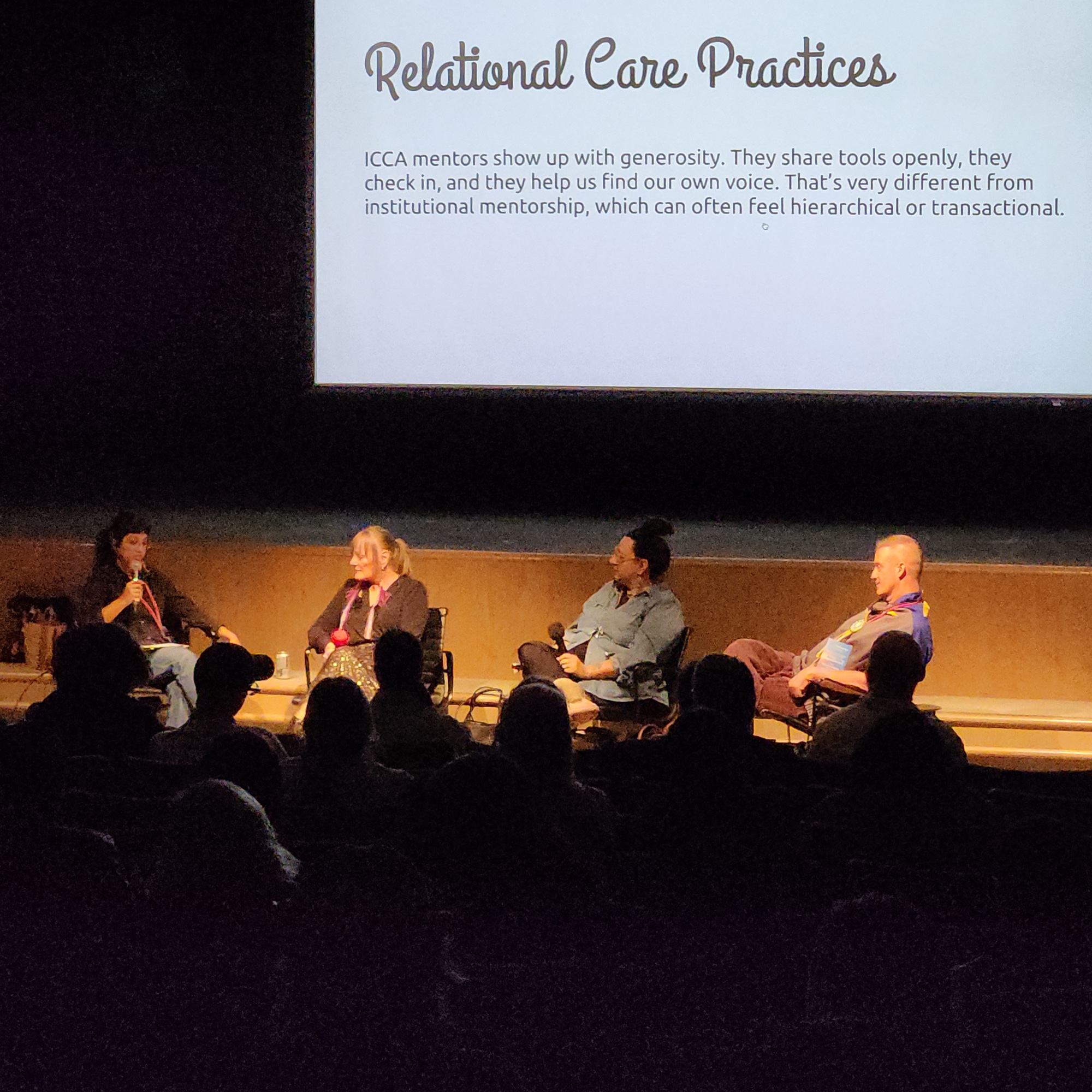Four panelists sit on a stage under a projected slide titled Relational Care Practices, as one speaks into a microphone. The audience sits in the dark in the foreground.