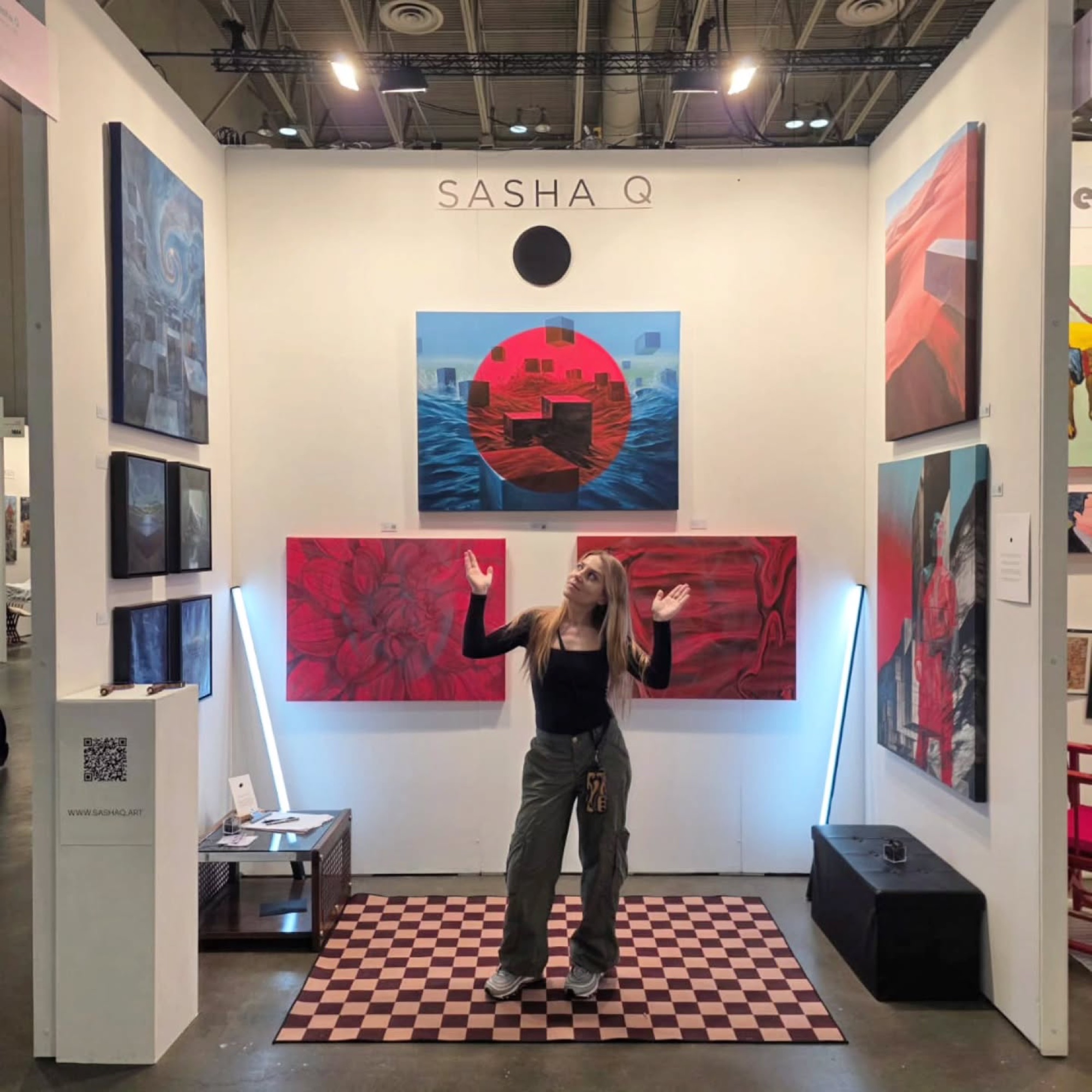 Sasha stands in her art fair booth under her name, with red and blue paintings and two light tubes.