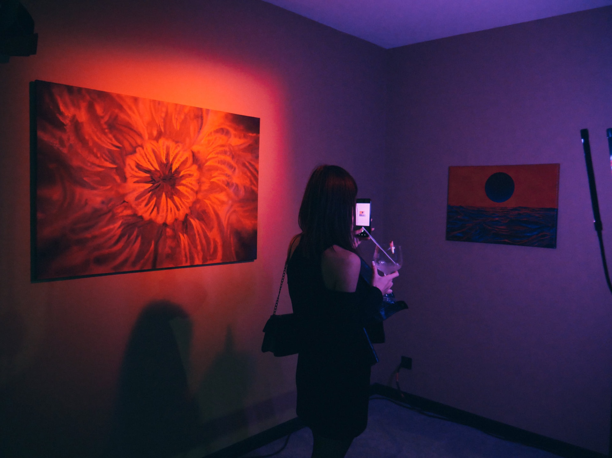 A visitor photographs a painting lit in red, in a dark room washed with purple light.