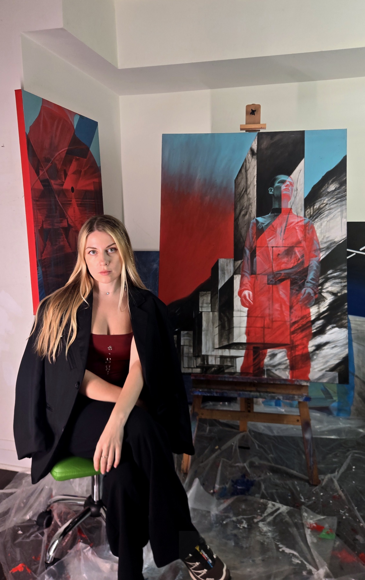 Sasha sits in her studio in front of two red and blue paintings, one on an easel.