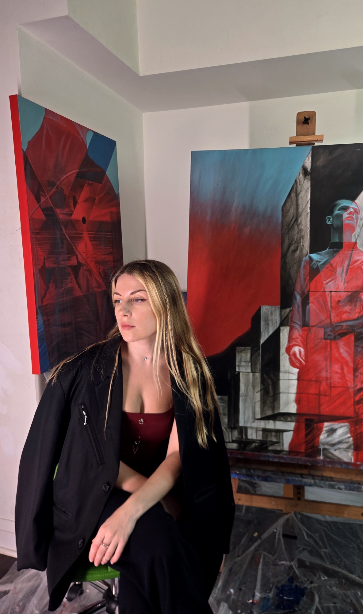 Sasha looks to the side in her studio, with a painting of a figure in red on the easel behind her.