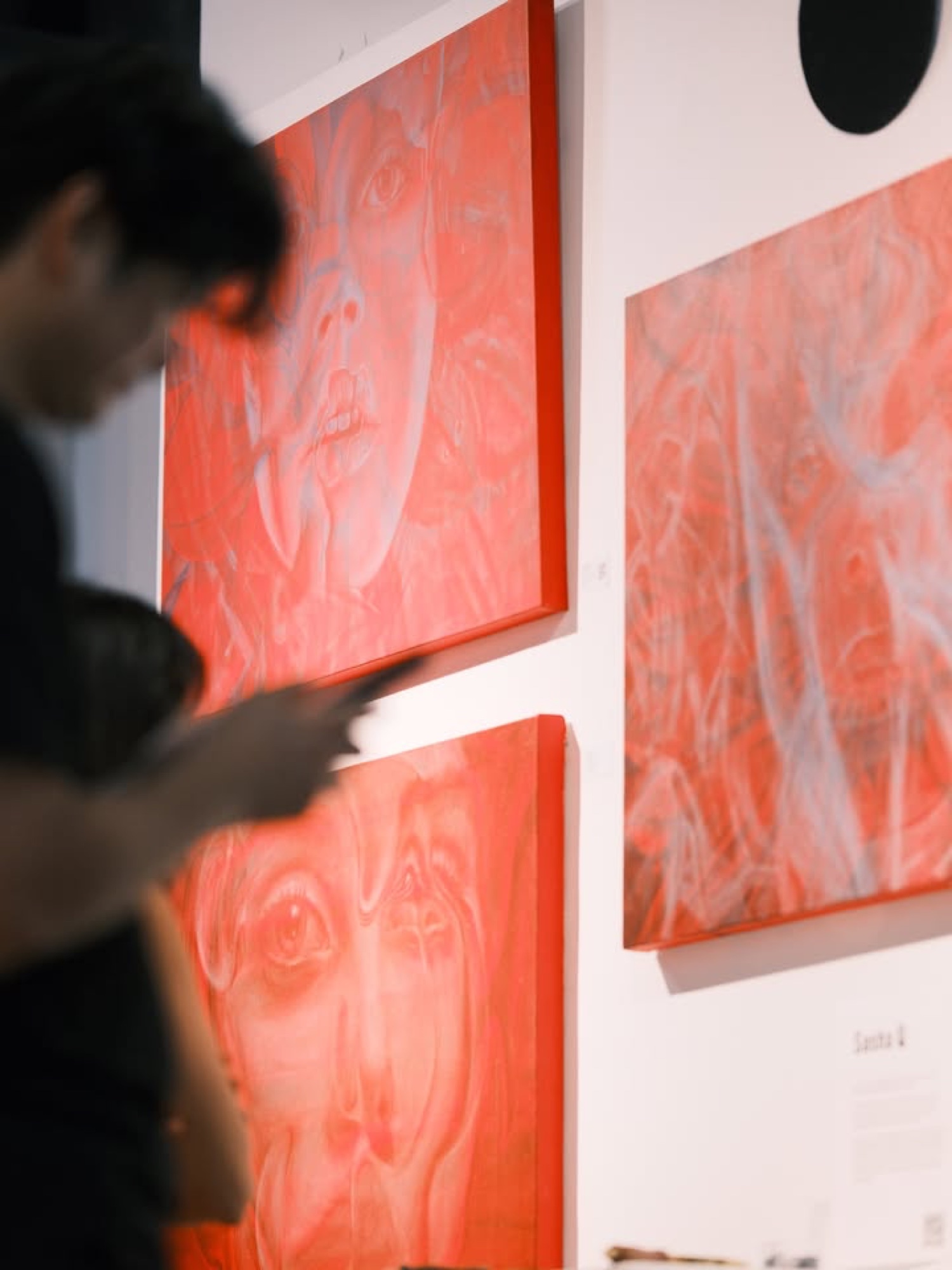 A visitor passes three red paintings of faces on a gallery wall.