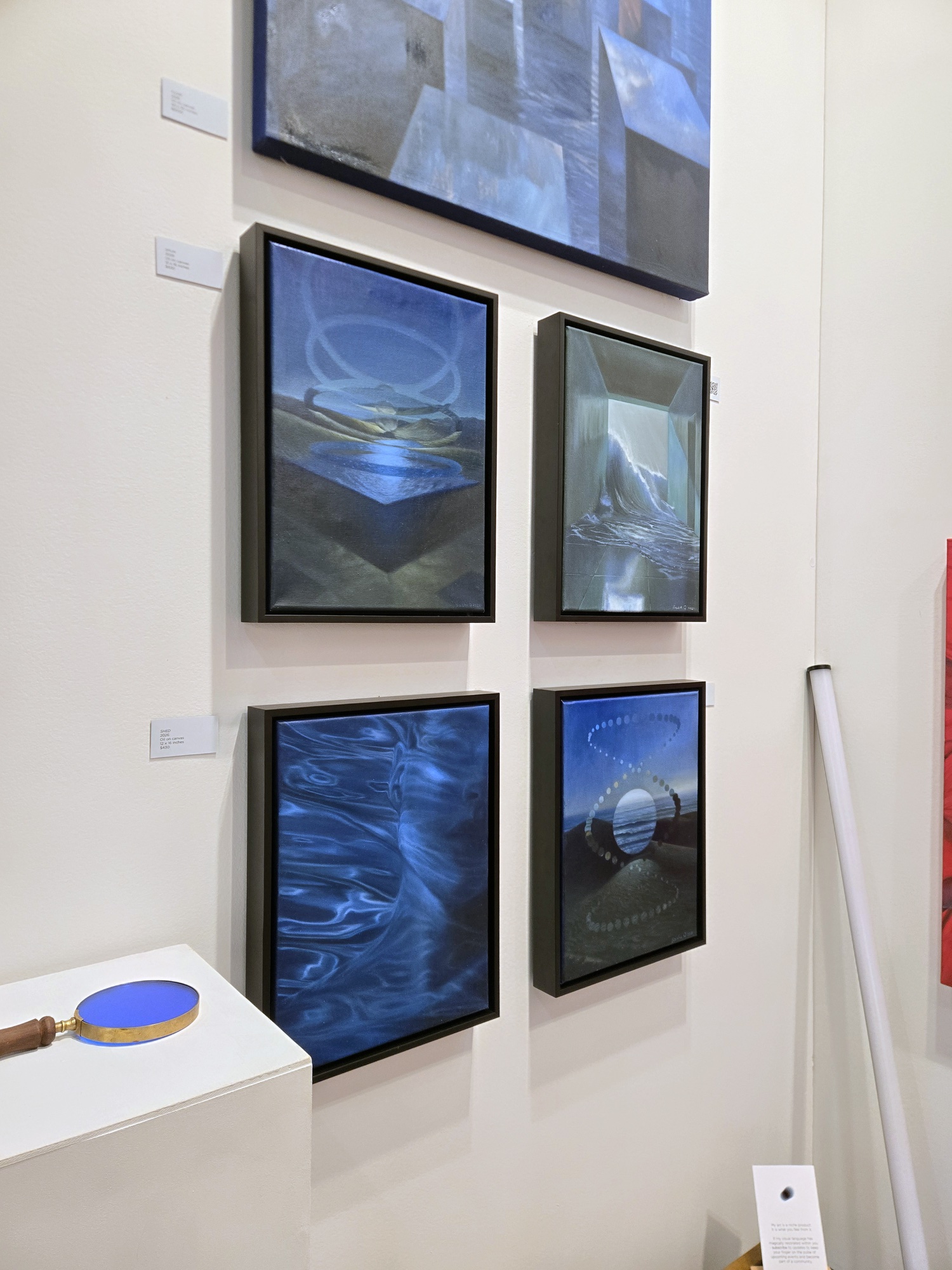 Four small framed blue paintings hang on a wall, with a blue magnifying glass on a plinth below.