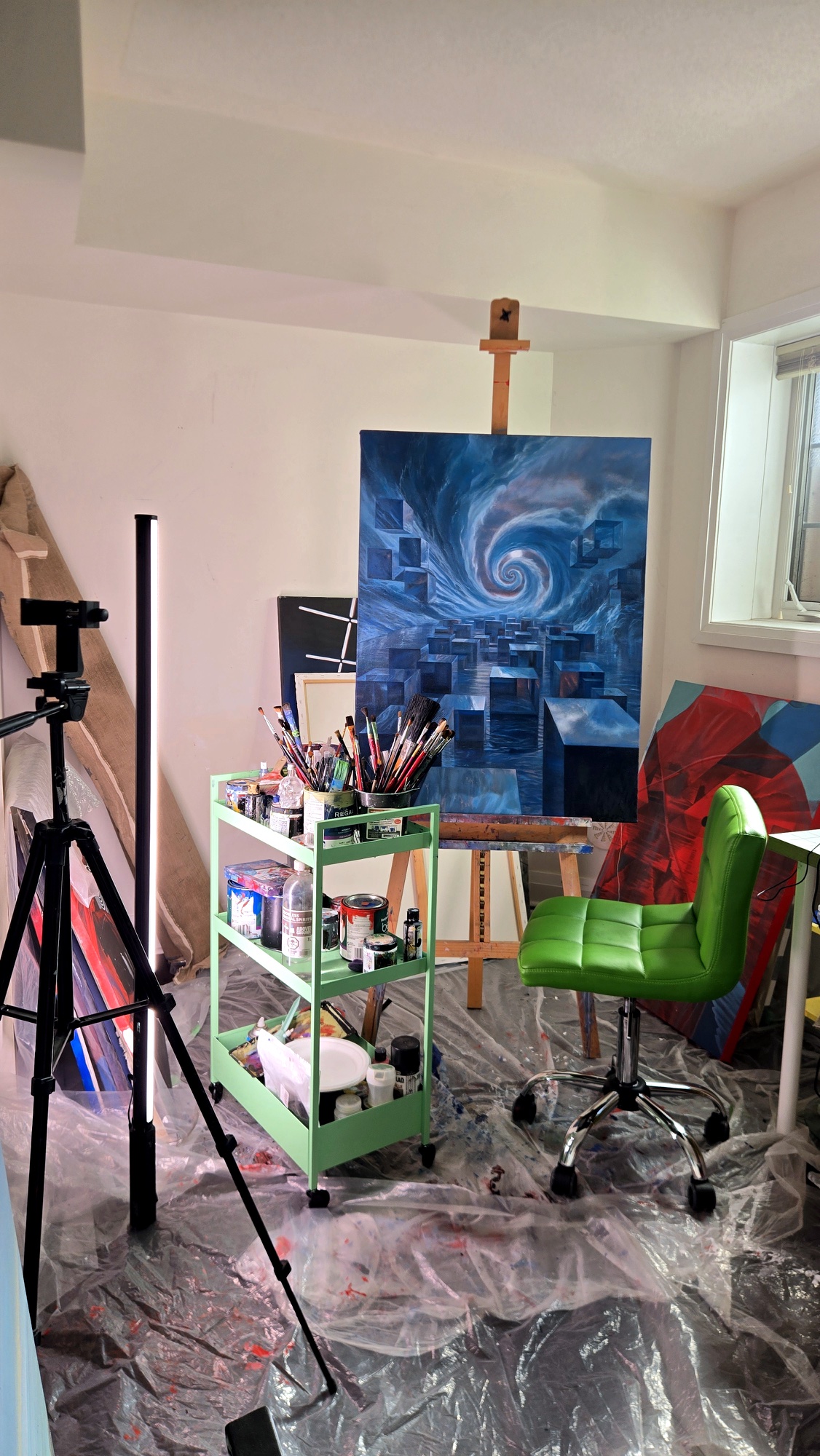A blue painting of a spiral sky over floating cubes stands on an easel beside a green cart of brushes.