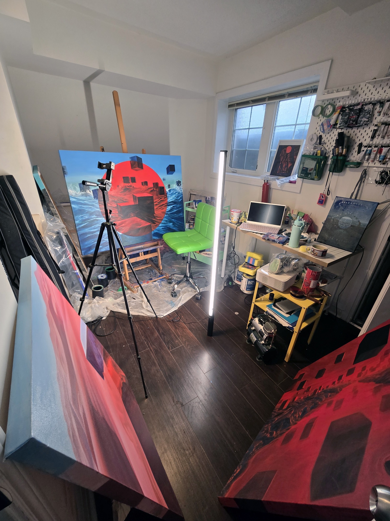 Sasha's studio, with a painting of a red sun and cubes over waves on the easel, a camera tripod and a light tube.